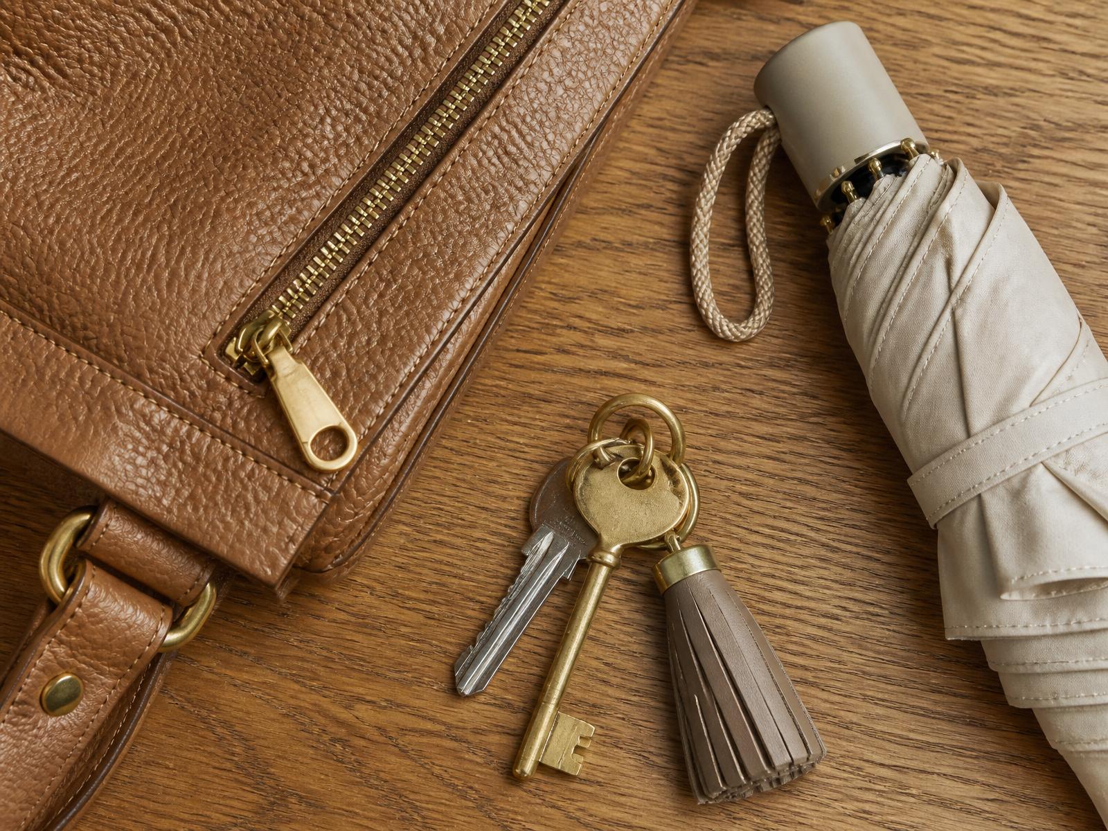 Detail of a pebbled tan leather bag with polished brass zipper, keys, and a folded umbrella on oak